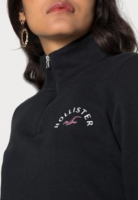 Kvinna som bär en svart zip-up-tröja med hög krage som har vit "Hollister"-text och en rosa mås-logotyp på bröstet, samt gyllene geometriska örhängen.