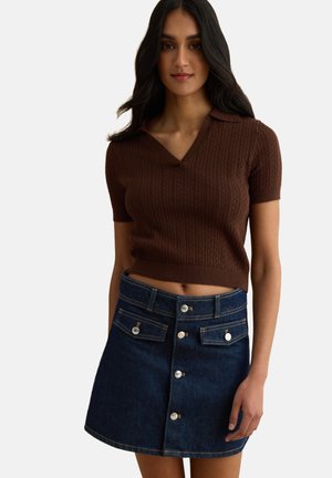Femme portant un haut en tricot marron à manches courtes et une jupe en jean bleu foncé à boutons sur le devant, debout devant un fond uni.