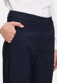 Pantaloni tailored di un blu navy scuro con una texture liscia, una tasca laterale e chiusura con bottone in vita. Design semplice e pulito.