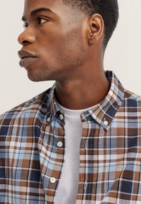 Braun und blau kariertes Hemd mit Button-Down-Kragen, ausgestattet mit weißen Knöpfen und einer leichten Stofftextur. Weißes Unterhemd ist darunter sichtbar.