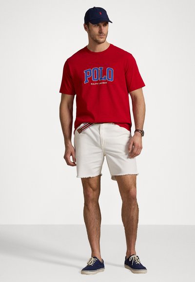 Red cotton t-shirt with "POLO" logo, white denim shorts with frayed hem, and navy cap, paired with navy sneakers and a multicolored belt.