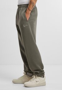 Olive green sweatpants with an elastic waistband, side pockets, and a small white logo on the thigh, paired with cream sneakers.