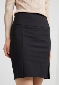 Jupe crayon noire avec une taille ajustée, texture lisse, détail de fente sur le côté et lignes épurées, associée à un haut beige à manches longues en côte.
