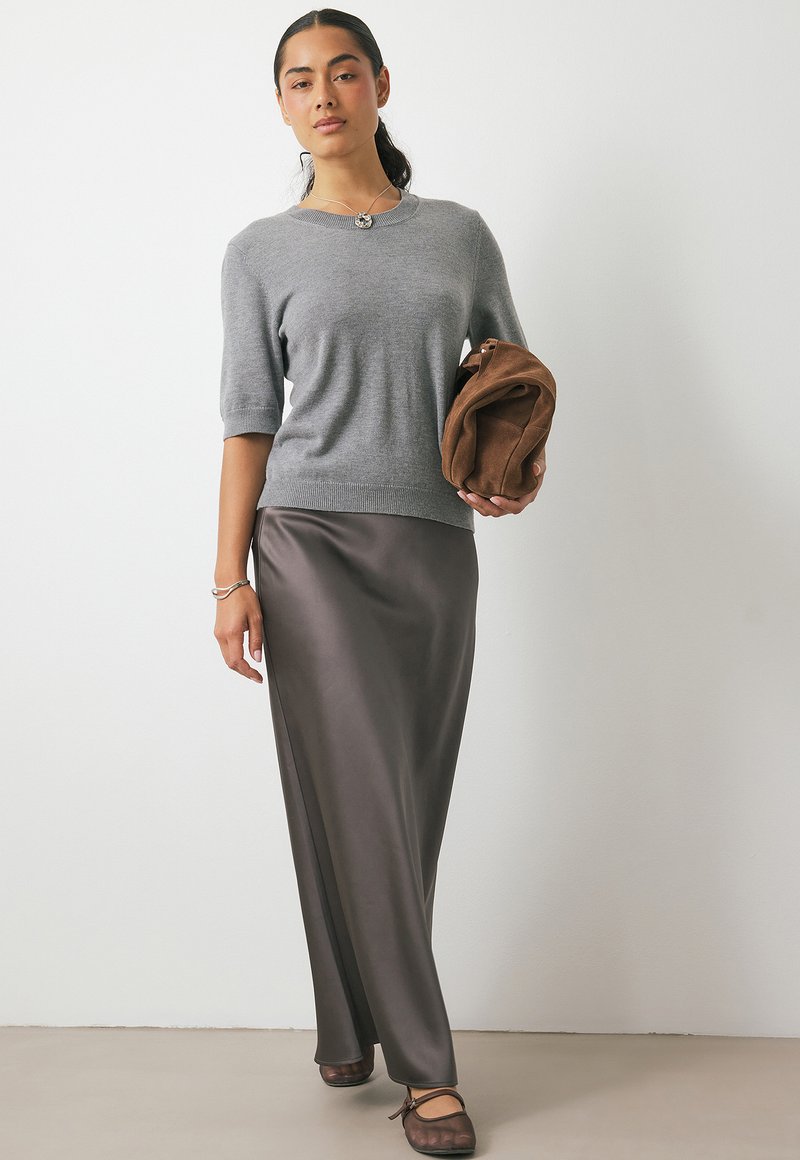 Donna che indossa un maglione grigio a maniche corte, una gonna maxi in raso scuro, scarpe scure, tiene in mano una clutch in suede marrone, in piedi contro un muro bianco.