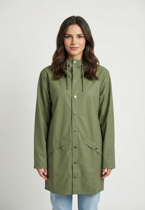 Femme aux cheveux mi-longs et foncés, portant un imperméable vert boutonné avec des poches et un jean bleu clair, debout devant un fond uni.