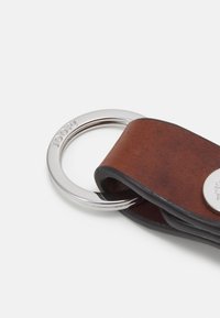 Porte-clés en cuir brun avec un anneau en métal argenté. Présente une finition texturée et un accent de logo circulaire, maintenu par une sangle noire.