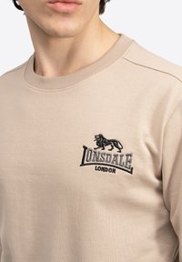 Bež jakna od mekog materijala sa okruglim izrezom, šavovima na ramenima i izvezenim crnim logotipom lava sa tekstom "Lonsdale London".