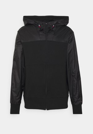 Veste à capuche noire zippée avec un design en tissu mixte comportant des manches lisses et un corps texturé, ainsi que des cordons sur la capuche.