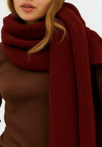 Kvinde iført et tykt, mørkerødt strikscarf viklet omkring halsen over en brun ribbet langærmet top med løst blond hår.