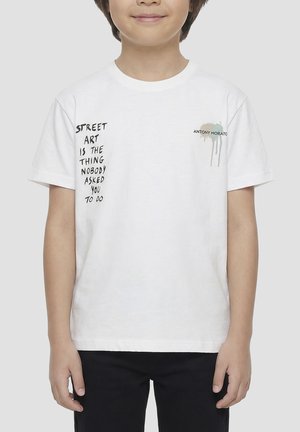 Enfant portant un t-shirt blanc avec un texte noir « Street art is the thing nobody asked you to do » et un petit logo tache de peinture colorée.