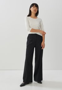 Weißes Langarmhemd kombiniert mit schwarzen, weiten Hosen aus glattem Stoff. Schwarze flache Schuhe runden das Outfit ab. Minimalistisches Design, keine Muster.