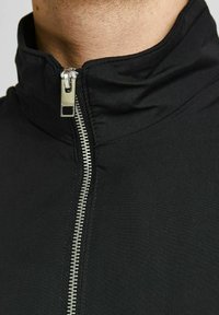 Veste noire avec un col montant et une fermeture éclair argentée visible. Le tissu semble lisse et mat avec un design structuré.