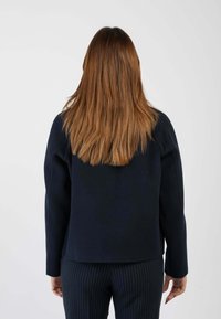 Giacca blu navy a maniche lunghe realizzata in un tessuto liscio, caratterizzata da un taglio dritto e un design minimalista, abbinata a pantaloni a righe verticali.