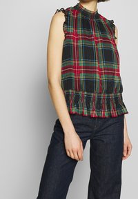 Débardeur à carreaux noir et rouge sans manches avec des accents de manches froncés, un décolleté froncé et un ourlet évasé et volanté. Porté avec un jean en denim foncé.