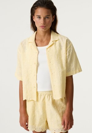 Kortärmelig lichtgele blouse met bloemrijke borduurwerk en open voorkant. Gecombineerd met shorts met scallopzoom in bijpassende stof.