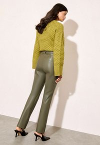 Olīvu zaļas pieguļošas bikses, kas saskaņotas ar teksturētu, adītu laima zaļu džempers. Melni papēžu mūļi noslēdz apģērbu.