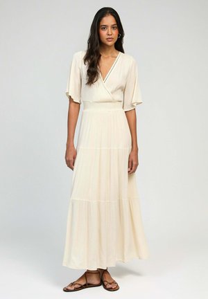 Robe maxi de couleur crème avec un style enveloppant, des manches courtes, un tissu texturé et un design à volants. Portée avec des sandales plates à lanières.