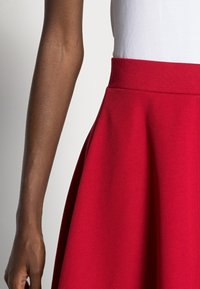 Jupe rouge évasée avec une texture lisse, dotée d'une taille ajustée et d'un drapé doux. Associée à un haut blanc, partiellement visible.