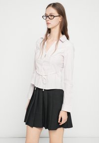 Πουκάμισο με λευκές και ροζ ρίγες, κουμπωτό, με λεπτομέρεια ζώνης στη μέση, συνδυασμένο με μαύρη πλισέ φούστα. Το ύφασμα φαίνεται απαλό και ελαφρύ.