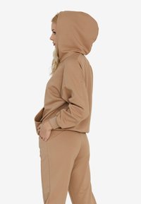 Beżowa bluza z kapturem, z prążkowanymi mankietami i przednią kieszenią, zestawiona z pasującymi dresami. Miękki materiał z luźnym krojem.