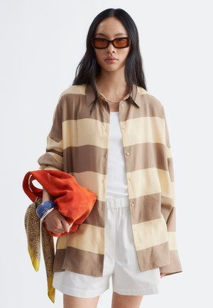 Femme aux longs cheveux noirs portant une chemise rayée marron et beige, des shorts blancs, des lunettes de soleil, tenant un sac orange avec une écharpe jaune et un bracelet bleu.