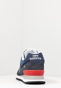 Zadní pohled na námořnickou modrou a šedou tenisku New Balance s červeným detailem na patě a bílým logem na bílé ploše a pozadí.