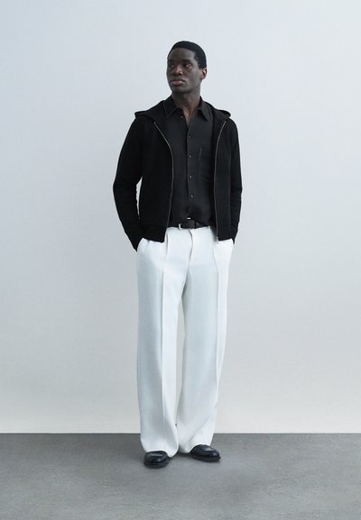 Άνδρας στέκεται με τα χέρια στις τσέπες, φορώντας μαύρο πουκάμισο, μαύρο ζακέτα με φερμουάρ, λευκό παντελόνι με φαρδιές πανκέτες και μαύρα παπούτσια, πάνω σε απλό φόντο.