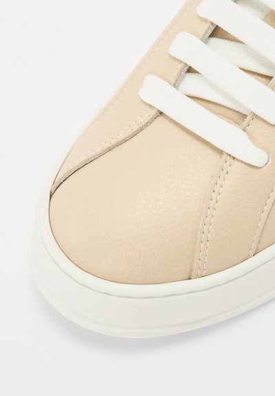 Beige leather sneaker featuring a textured surface, white flat laces, and a white rubber sole. The design includes stitched accents.