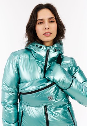 Žena u sjajnoj tirkiznoj jakni s punjenjem i odgovarajućoj torbici za struk, drži ovratnik jakne, neutralan izraz lica, bijela pozadina.