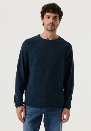 Mann mit kurzen lockigen Haaren, der einen dunkelblauen Pullover mit langen Ärmeln und blaue Jeans trägt, steht vor einem einfarbigen hellen Hintergrund.