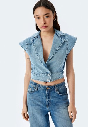 Chaqueta de mezclilla recortada con hombreras, de color azul claro, escote en V profundo y cierre de cinco botones, combinada con jeans azul de cintura alta.