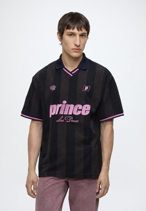 Young man with short brown hair wearing a black and dark striped polo shirt with pink text "prince Les Deux" and pink-trimmed sleeves.