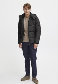 Sort fiberjakke med hætte, lynlås foran og sidelommer, over en lys beige sweater, parret med mørkeblå jeans og grå sko.