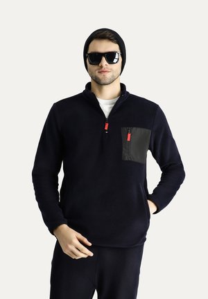 Pullover in pile blu navy con tasca sul petto in nylon nero, tirazip rossi, colletto alto e maniche lunghe. Indossato con occhiali da sole e un berretto.