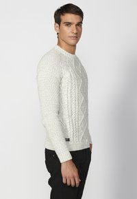 Chunky weißer Kabelstrickpullover mit gerippten Bündchen und Saum, verfügt über einen runden Halsausschnitt. Der Model steht seitlich in schwarzen Hosen.