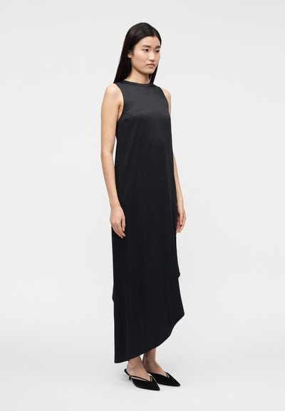 Vestido preto sem mangas com textura suave, bainha assimétrica e design minimalista. Combinado com sapatilhas pretas pontiagudas.