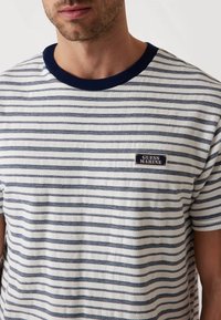 Camiseta a rayas en blanco y azul marino, con un cuello de canalé azul marino y una pequeña etiqueta "Guess Marine" en la parte delantera. Textura de algodón suave.