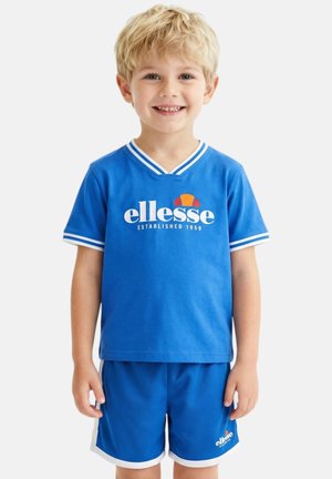 Jeune garçon aux cheveux blonds portant une tenue de sport bleue ellesse, souriant, debout devant un fond blanc uni.