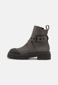 Šedá nubuková kotníková bota s gumovou špičkou, texturovanou podrážkou a nastavitelným popruhem s detailem sponky. Obsahuje poutko vzadu.