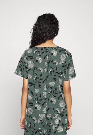 Haut floral vert avec des manches courtes et un col rond. Le tissu a une apparence texturée avec des motifs floraux blancs et noirs.