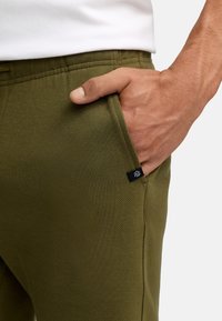 Mano descansando en el bolsillo frontal de pantalones texturizados verde oliva, combinados con una blusa blanca, mostrando el detalle de la tela y una pequeña etiqueta negra.