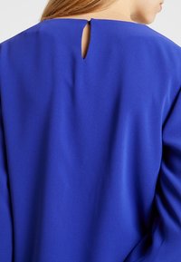Top blu con una finitura liscia e leggermente strutturata. Caratterizzato da un'apertura a chiave sul retro e maniche larghe e fluenti.