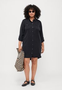 Black button-up dress with two chest pockets, rolled sleeves, and a slightly curved hem. Person holds a striped tote bag and wears black flats.