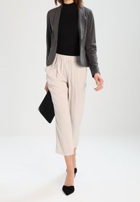 Kvinde-model iført en sort turtleneck og grå cardigan over cremede, højtaljede bukser, holder en sort clutch og har sorte hæle på.