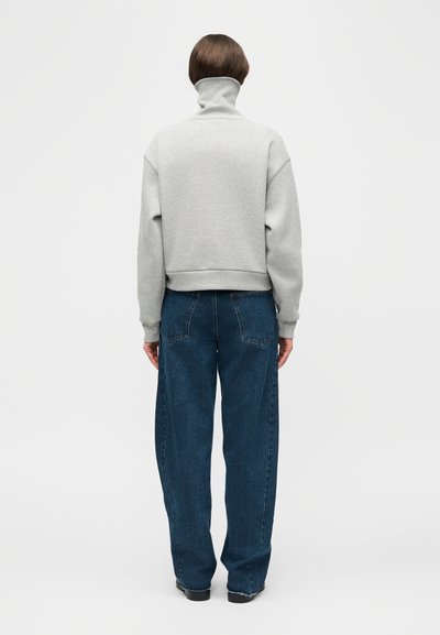 Άτομο με κοντά καστανά μαλλιά στέκεται με την πλάτη γυρισμένη φορώντας ανοιχτό γκρι πουλόβερ με ψηλό γιακά και φαρδιά σκούρα μπλε τζιν παντελόνια.
