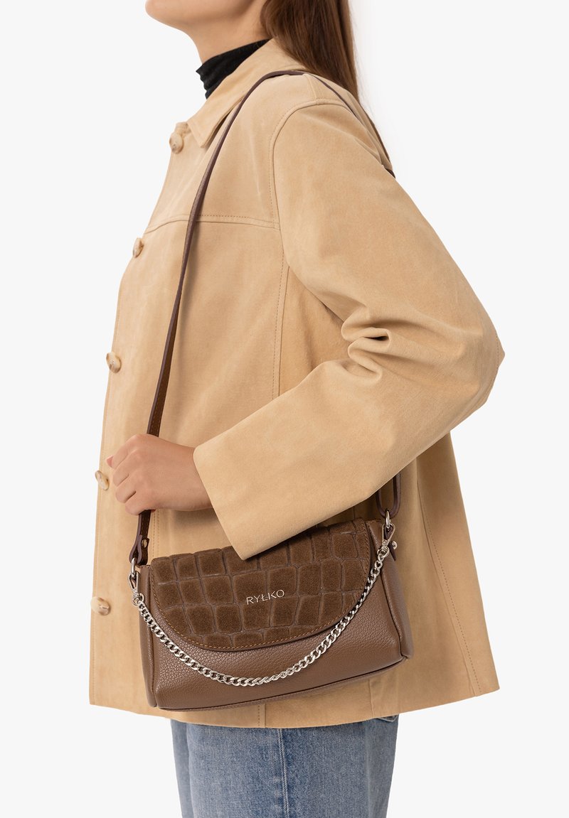 Femme portant un manteau beige et un jean bleu tenant un sac à main marron texturé avec une bandoulière en chaîne argentée sur l'épaule.