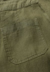 Vihreä oliivinvärinen pellavakangas sisältää yhden takataskun, joka on suorakaiteen muotoinen ja jossa on siistit ompeleet, mikä korostaa teksturoitua, luonnollista kudontaa.