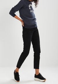 Jeans skinny neri con orlo grezzo, abbinati a una felpa blu navy con testo bianco. Sneakers nere con suola bianca completano il look.