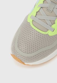 Ανοιχτό γκρι αθλητικό παπούτσι με αναπνεύσιμη επιφάνεια από διχτυωτό υλικό, πράσινες λεπτομέρειες και υφασμένη ελαστική σόλα με αντίθετο ταμπά πέλμα.