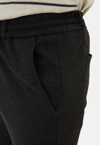 Černé kalhoty s texturou, s kónickým střihem, vybavené předními kapsami a vysokým pasem, částečně zakryté světle zbarveným topem.